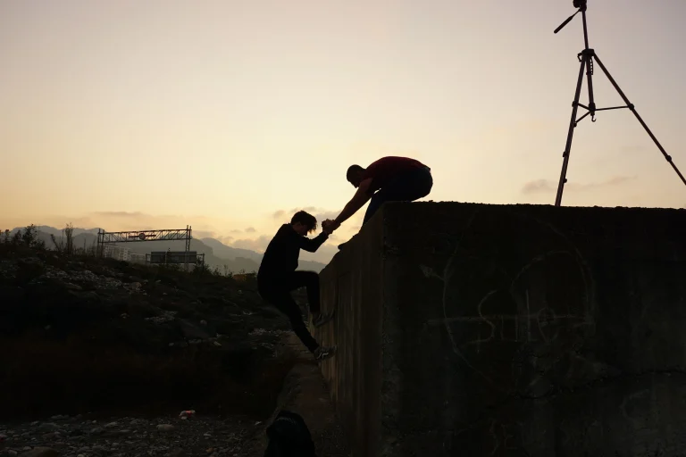 Silhouette of one man helping another climb up a wall at sunrise