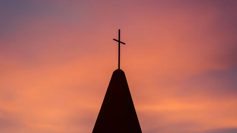 Silhouette of a cross on a church steeple against a glowing sunset sky