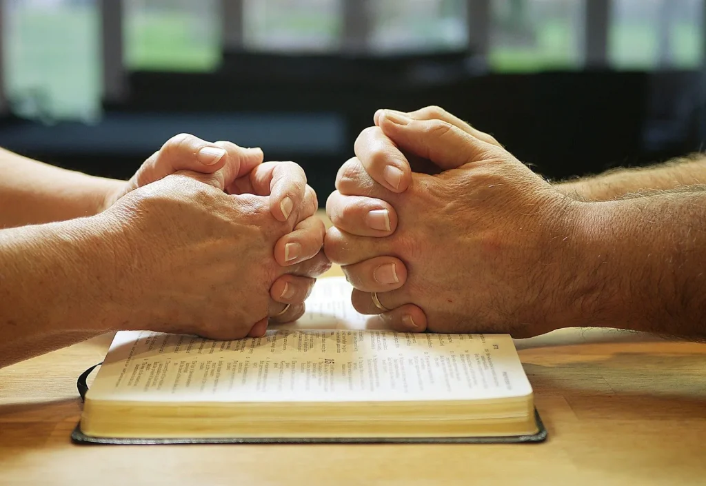 Two people holding hands over an open book as they pray together.
